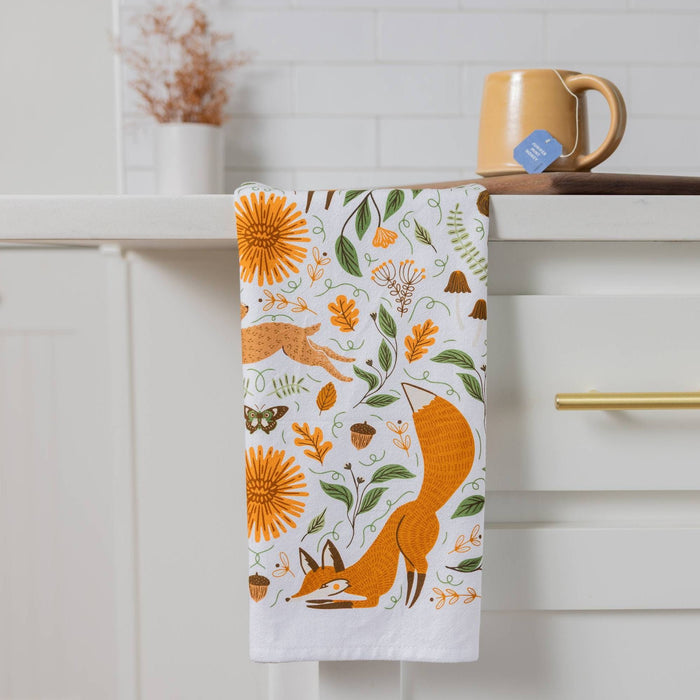 A white kitchen towel with a colorful autumn forest pattern featuring a fox, sunflowers, acorns, and leaves, hanging from a white cabinet handle in a kitchen setting.