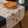 A rectangular table runner with an orange, green, and white floral pattern draped over a dark table, styled with plates and folded napkins.