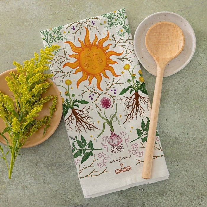 Tea towel with sun and plant design, wooden spoon, and bowl of yellow flowers on a green surface