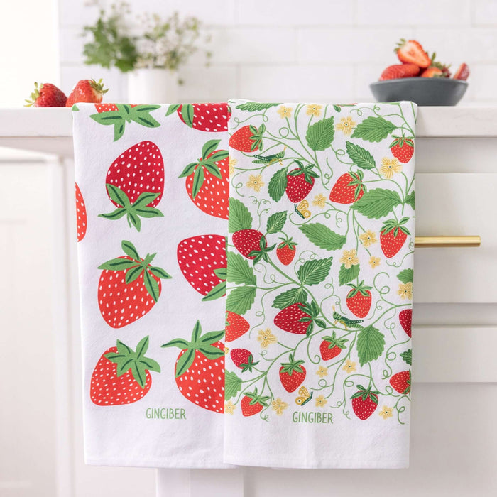Two strawberry-themed towels hanging on a rack with a kitchen setting in the background.